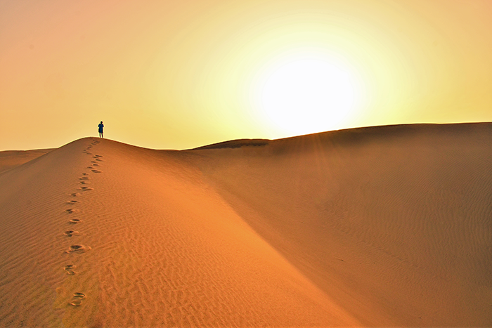 Photo of a person walking in the desert, leaving foot tracks in the sand with a very bright sun in the background